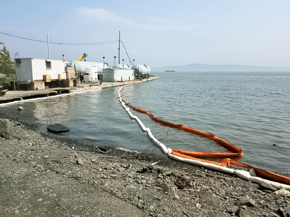 Absorbent Boom for Oil Spill Cleanup and Containment White absorbent boom deployed alongside an orange oil containment boom near an industrial shoreline to absorb oil and hydrocarbons from the water surface.