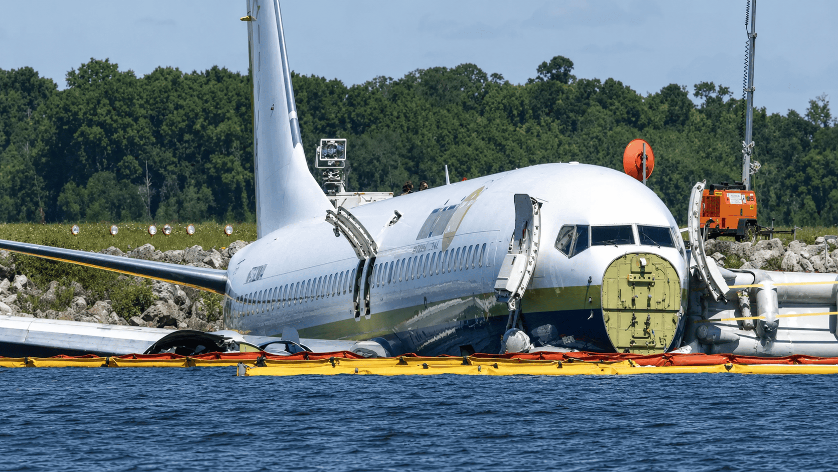 Oil Containment Boom for Emergency Fuel Spill Response Yellow and orange oil containment boom deployed around a damaged aircraft near water to help contain fuel, oil, and other floating hydrocarbons during emergency spill response.